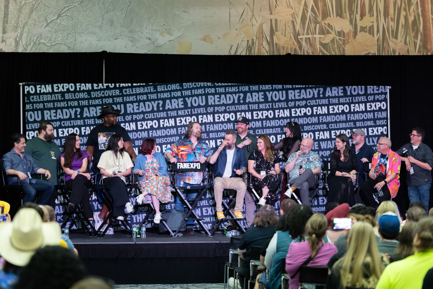 A wide panel shot of over a dozen One Piece voice actors and creators seated in a row onstage at FAN EXPO, laughing and chatting in front of a branded backdrop that reads “ARE YOU READY?” and “DISCOVER. CELEBRATE. BELONG.”