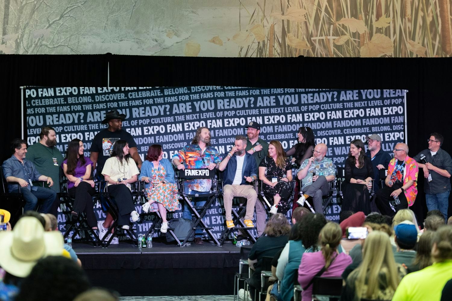 A wide panel shot of over a dozen One Piece voice actors and creators seated in a row onstage at FAN EXPO, laughing and chatting in front of a branded backdrop that reads “ARE YOU READY?” and “DISCOVER. CELEBRATE. BELONG.”