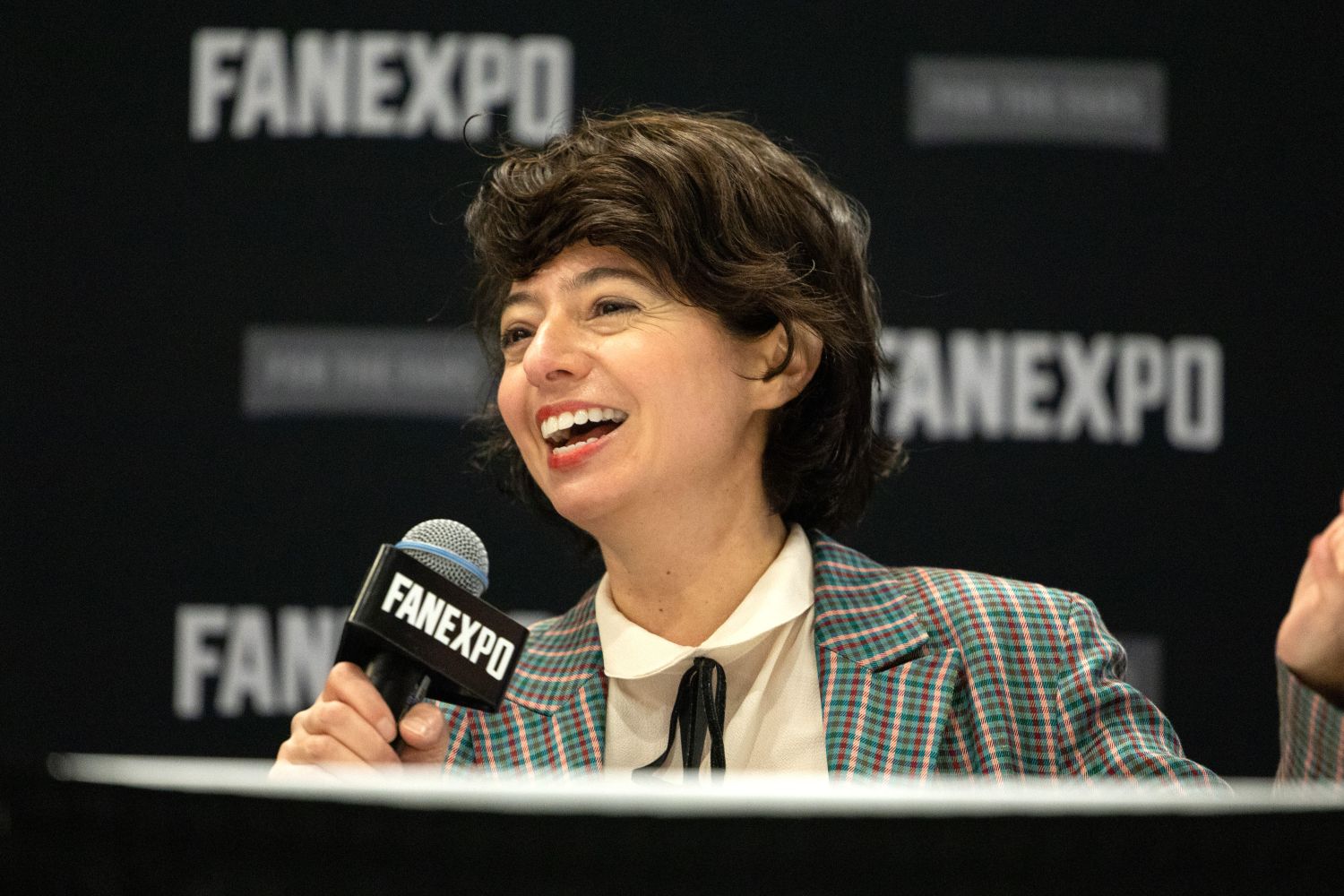 Kate Micucci, wearing a checkered blazer and black bowtie-style ribbon at the collar, laughs while speaking into a FAN EXPO mic at her panel table.