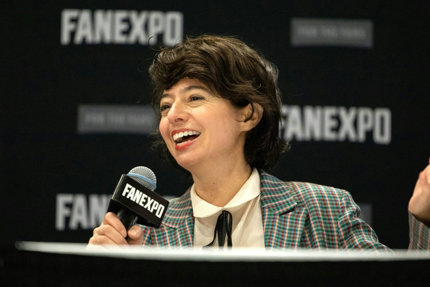 Kate Micucci, wearing a checkered blazer and black bowtie-style ribbon at the collar, laughs while speaking into a FAN EXPO mic at her panel table.