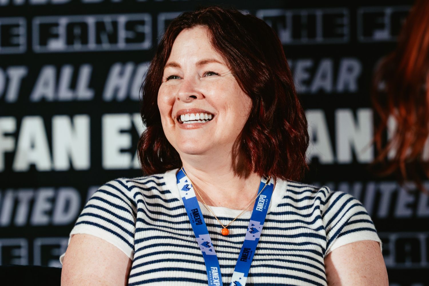 A close-up of Luci Christian smiling brightly onstage at FAN EXPO. She wears a striped shirt and blue event lanyard, with the blurred backdrop behind her featuring the FAN EXPO logo.