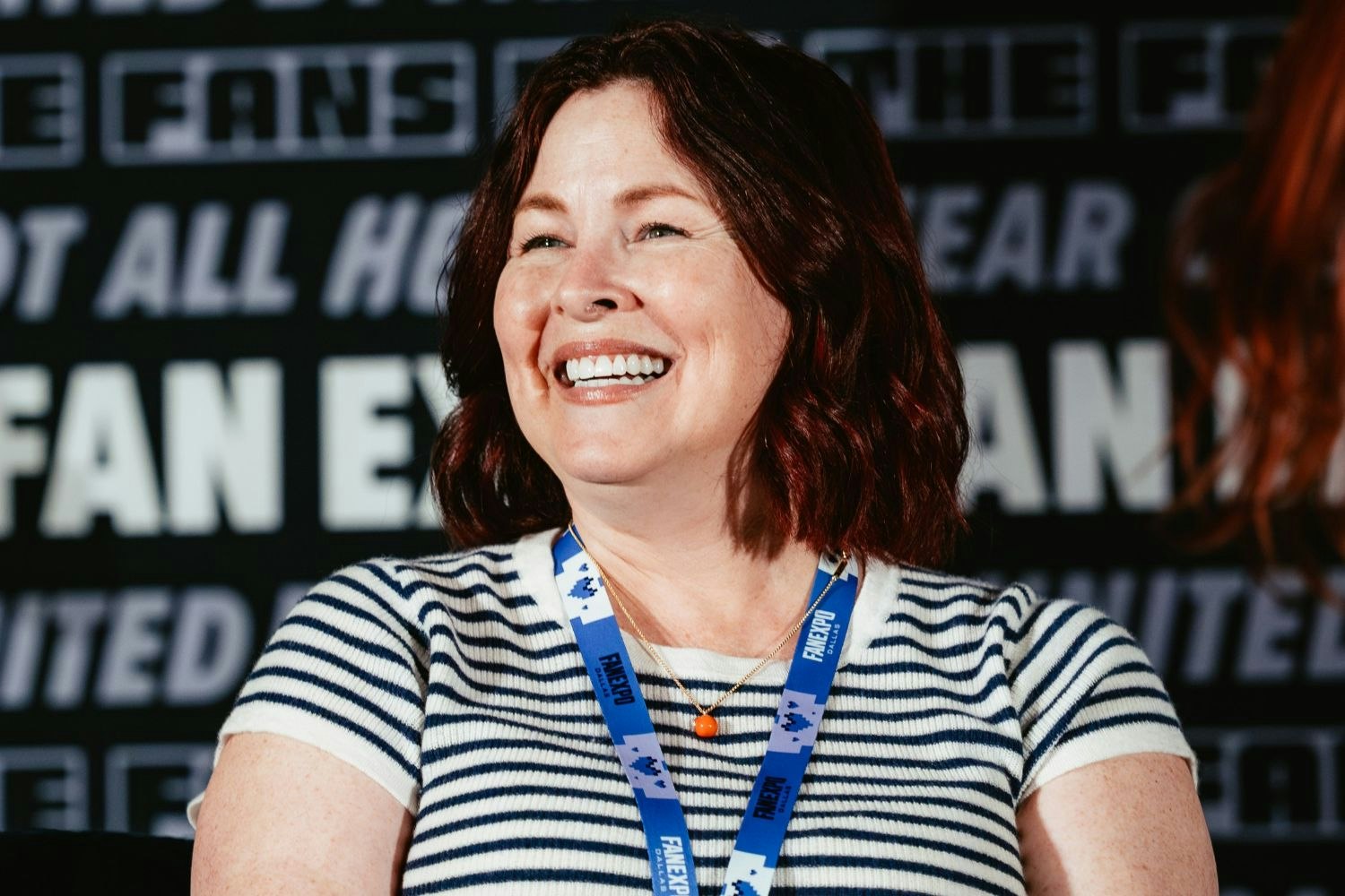 A close-up of Luci Christian smiling brightly onstage at FAN EXPO. She wears a striped shirt and blue event lanyard, with the blurred backdrop behind her featuring the FAN EXPO logo.