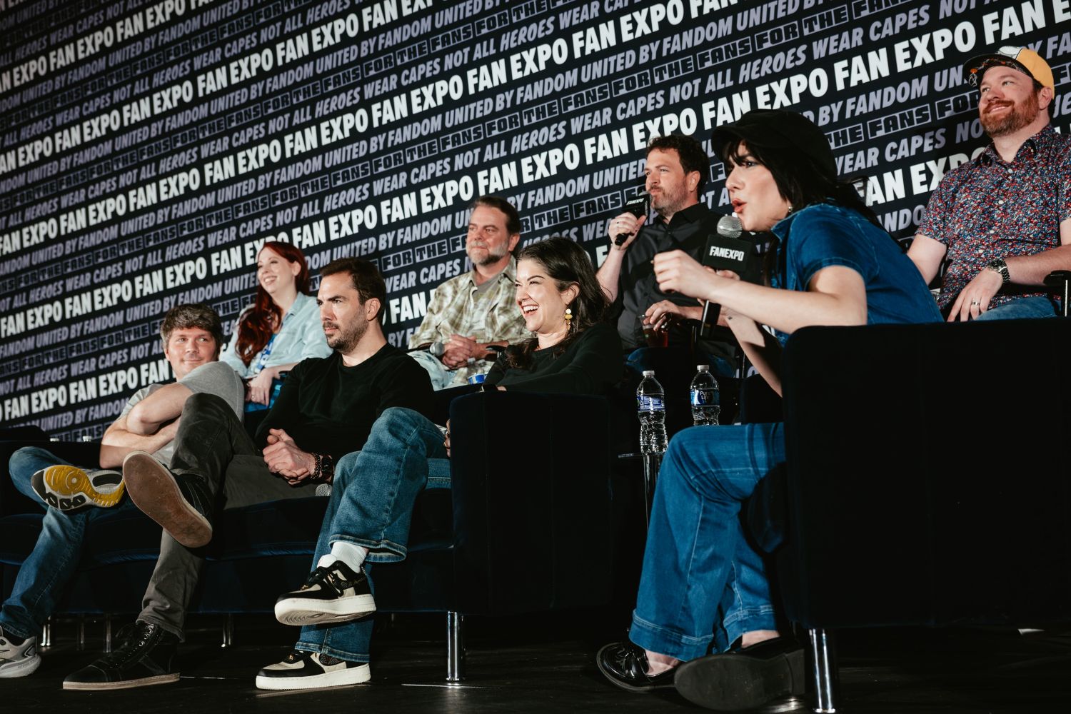 A group of nine voice actors and creators participate in a lively panel at FAN EXPO, seated on couches and chairs. One panelist leans forward to speak while others laugh and listen.