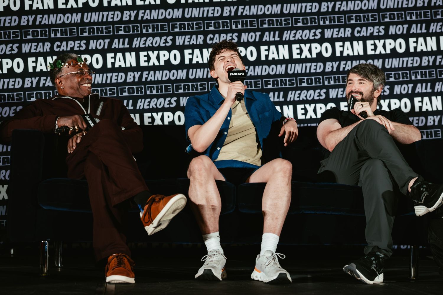 Three Hazbin Hotel voice actors on a couch at a FAN EXPO panel laugh together. Keith David wears a crown of vines, Blake Roman sits centerstage in shorts and a button-up shirt mid-laughter, while Alex Brightman smiles, holding a mic.