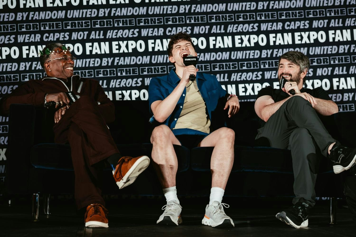 Three Hazbin Hotel voice actors on a couch at a FAN EXPO panel laugh together. Keith David wears a crown of vines, Blake Roman sits centerstage in shorts and a button-up shirt mid-laughter, while Alex Brightman smiles, holding a mic.