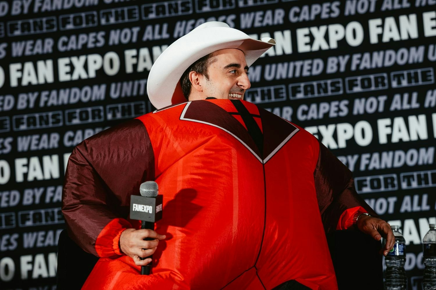 Amir Talai, dressed in an inflatable red and brown cowboy costume and white cowboy hat, laughs while holding a FAN EXPO mic onstage in front of a branded backdrop.