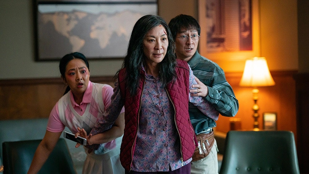 A screen cap from Everything Everywhere All At Once, featuring the three main characters of the film all standing together as they are surprised by an unscene character. From left to right: Joy Wang (played by Stephanie Hsu), Evelyn Wang (played by Michelle Yeoh), and Waymond Wang (played by Ke Huy Quan).