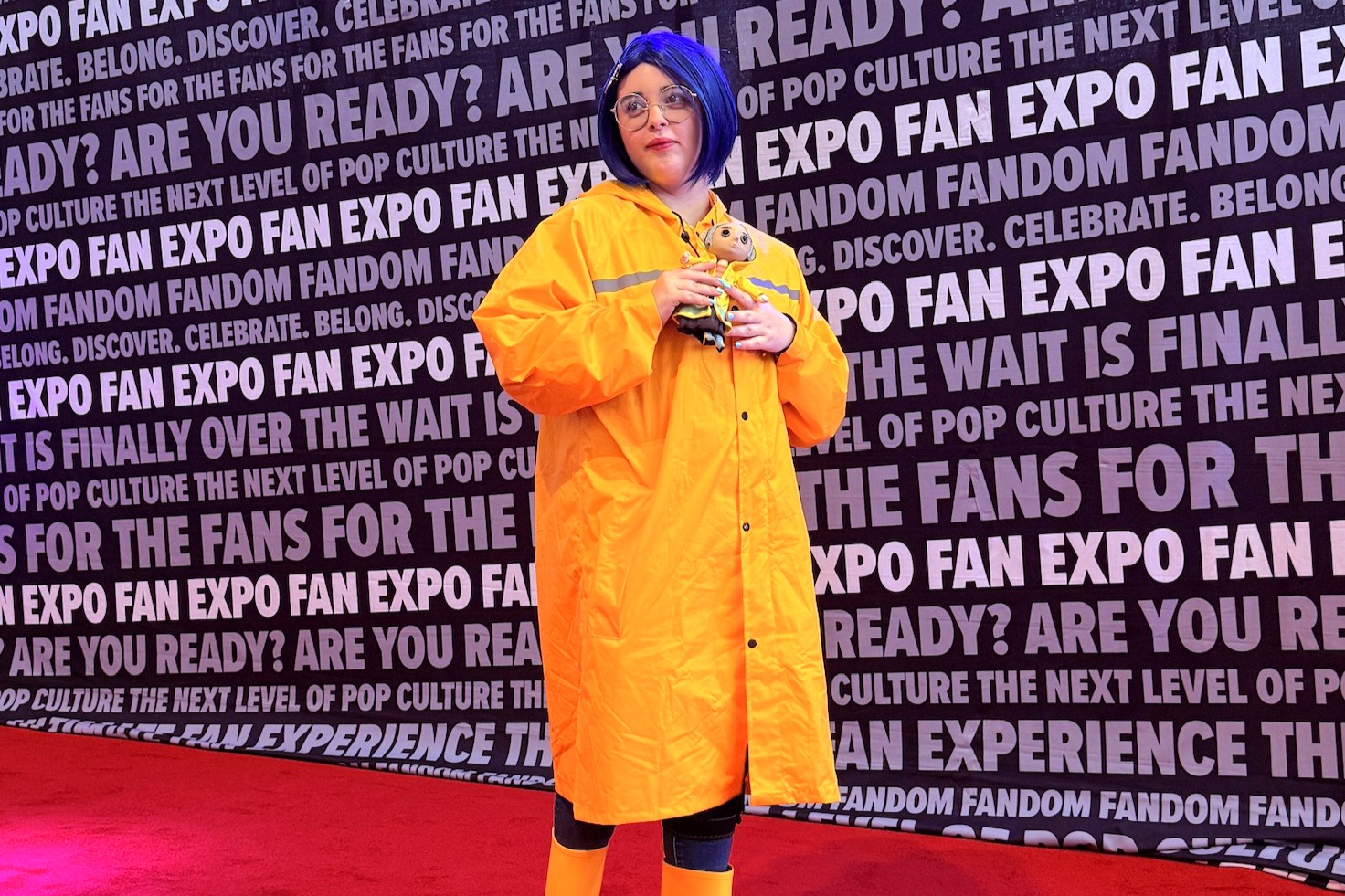 A cosplayer dressed as Coraline stands on the FAN EXPO red carpet wearing a bright yellow raincoat and blue wig, holding a Coraline doll and smiling slightly toward the camera.