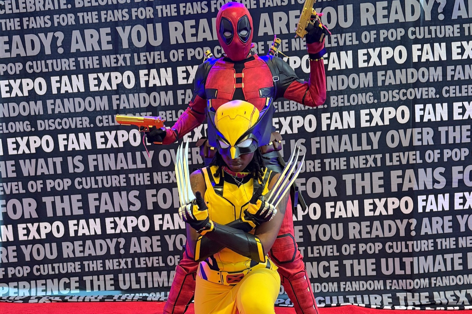 Two cosplayers in a dynamic pose on the red carpet portray Deadpool and Wolverine. Deadpool stands tall with toy pistols, while Wolverine crouches in front, claws out in a fierce stance.