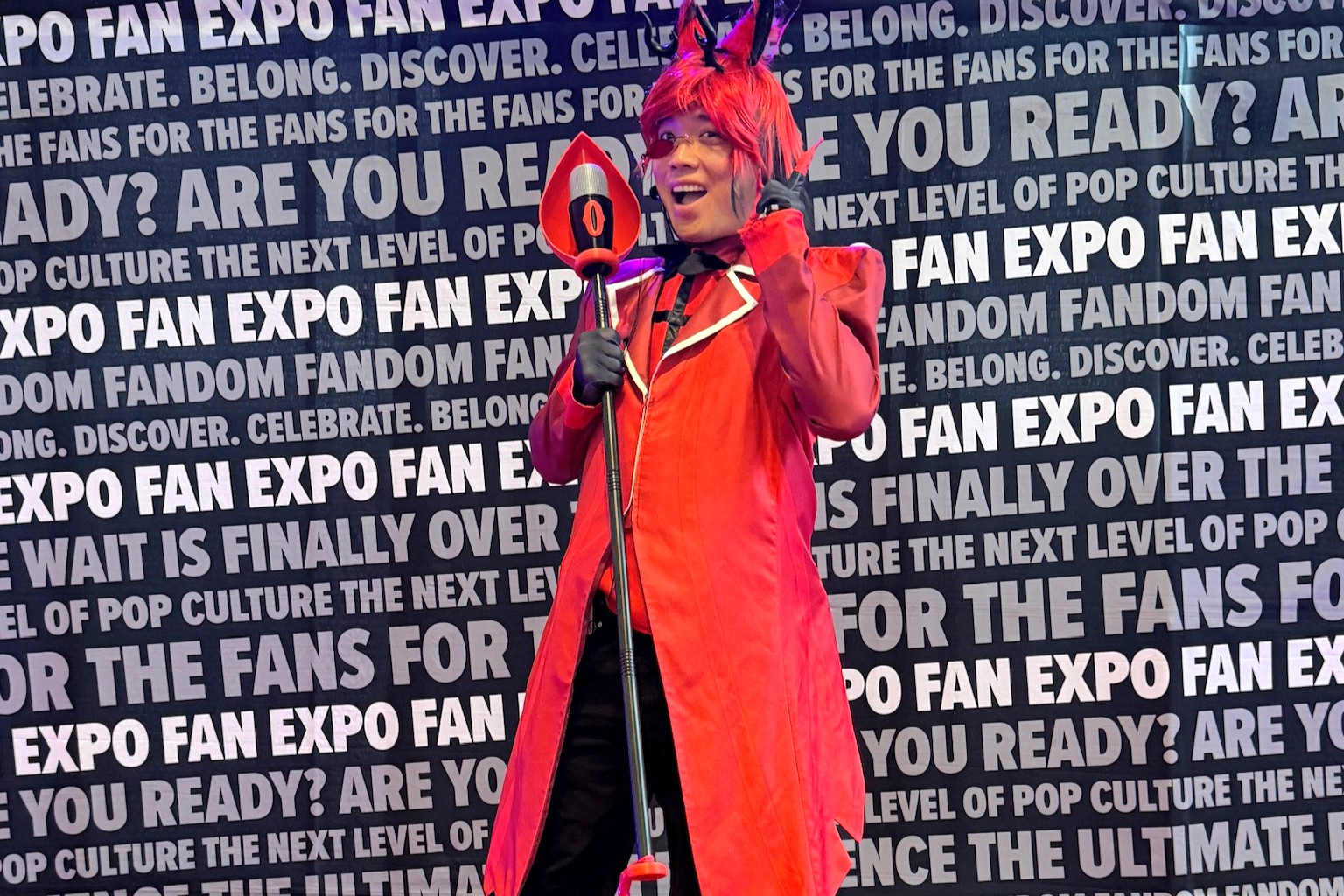 A cosplayer dressed as Alastor from Hazbin Hotel poses with a cheeky smile, wearing a red suit, red wig, black gloves, and holding the character’s iconic microphone staff.
