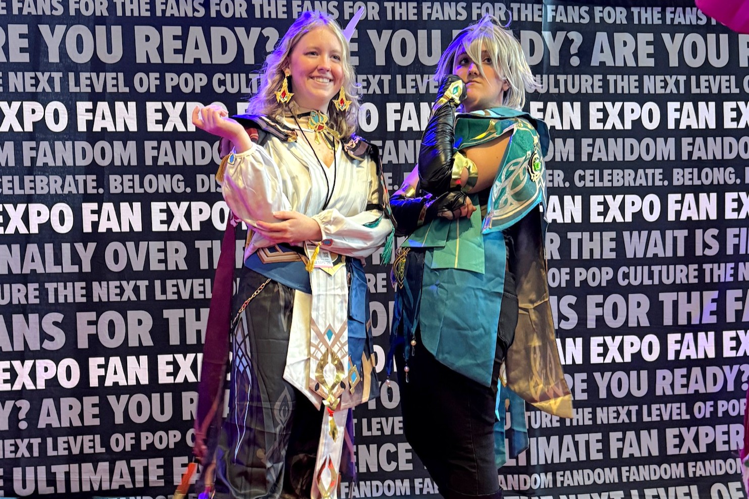 Two cosplayers dressed as characters from Genshin Impact pose proudly in detailed fantasy costumes featuring ornate embroidery, layered fabrics, and magical props.