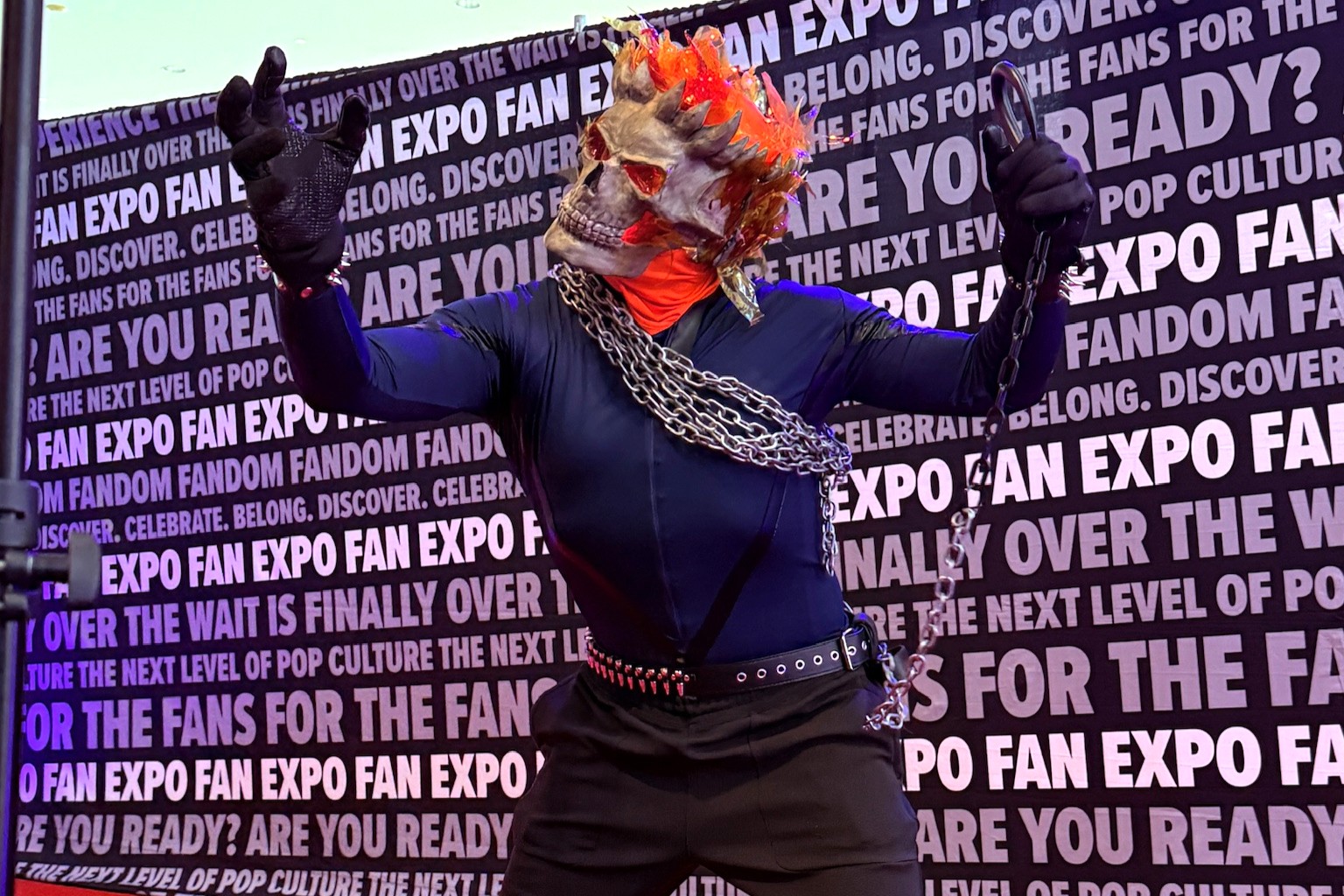 A cosplayer brings Ghost Rider to life with a skeletal flame helmet, black bodysuit, chains draped across their torso, and a fierce pose with outstretched arms on the red carpet.
