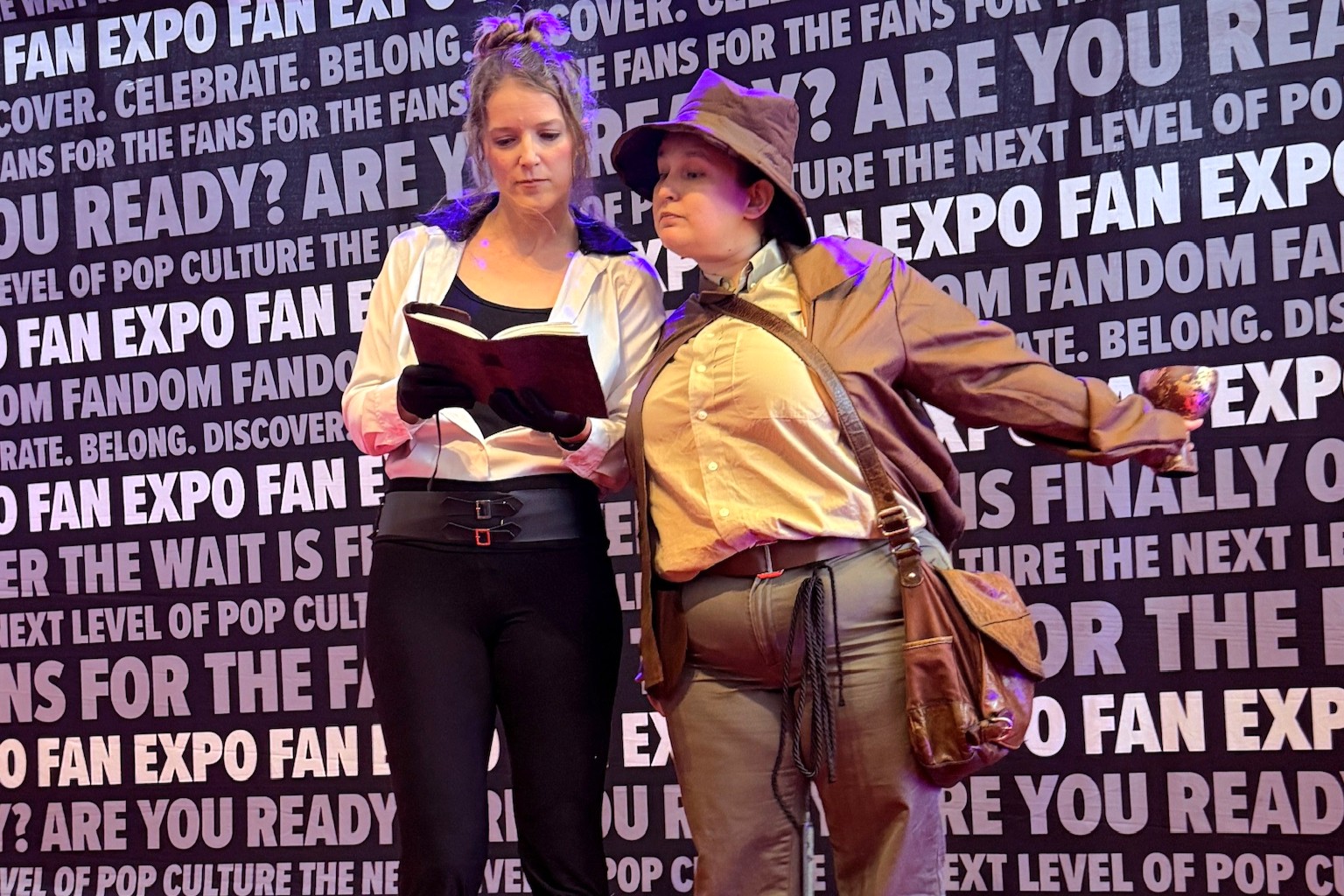 Two cosplayers channel Indiana Jones and Elsa Schneider, with one holding a prop grail and wearing a brown fedora and satchel, while the other studies a leather-bound journal in a white shirt and black pants.