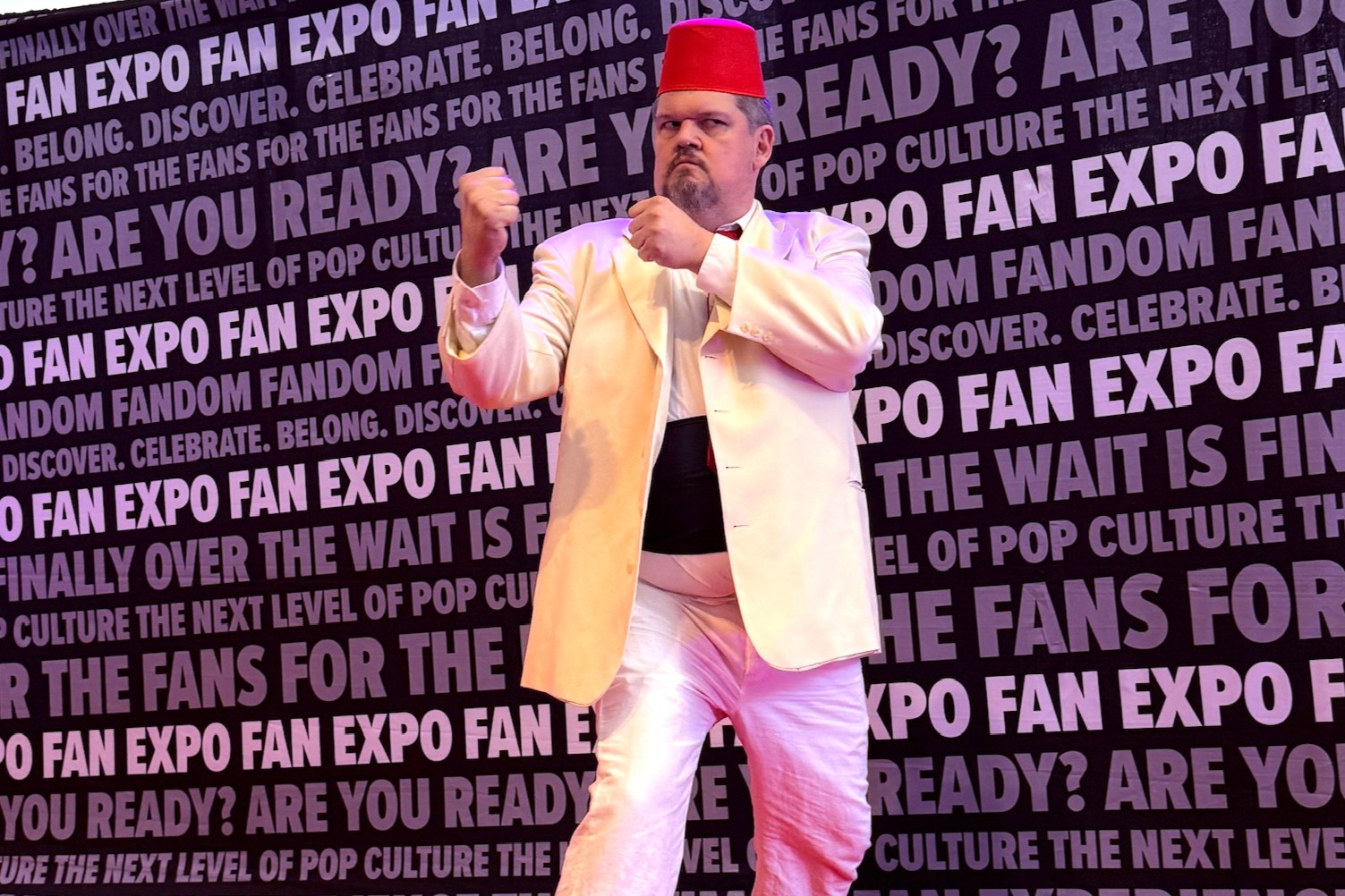 A cosplayer dressed in a striking white suit and red fez hat poses on the red carpet in a fighting stance, evoking the look of a classic villain or vintage film character.