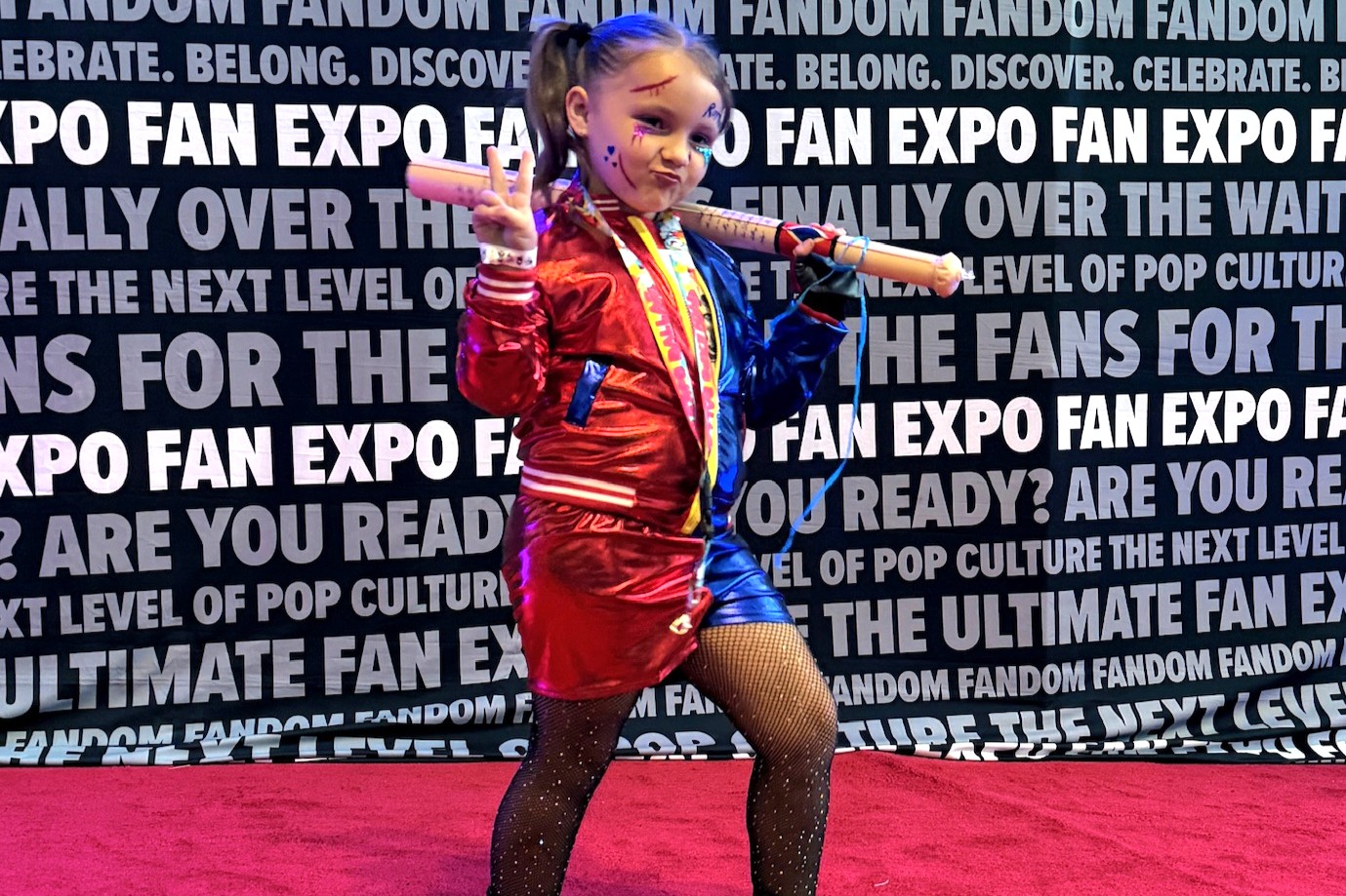A young cosplayer dressed as Harley Quinn from Suicide Squad throws up a peace sign while holding a bat over her shoulders, rocking a split red and blue outfit with fishnet stockings and face paint.