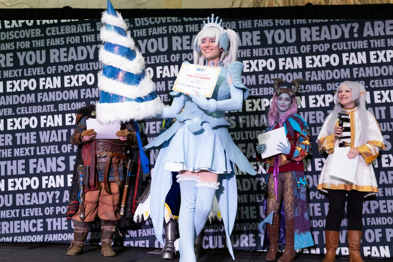 A smiling cosplayer in a pale blue fantasy armor set poses mid-stage holding a “Judge’s Award” certificate. They wield a massive, spiraling blue and white cone-shaped weapon. Behind them, other colorful contestants—including a tiefling with horns—watch and applaud.