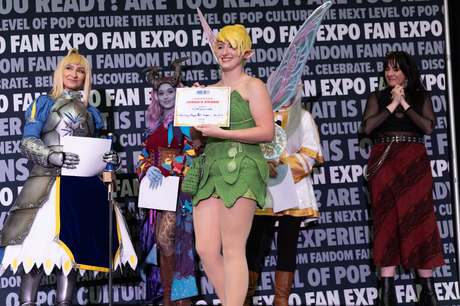A cosplayer dressed as a handmade fairy in a crochet-style Tinker Bell outfit smiles brightly while holding a “Judge’s Award” certificate. Their shimmering wings and pixie haircut complete the whimsical look. Other fantasy-clad finalists cheer behind them.