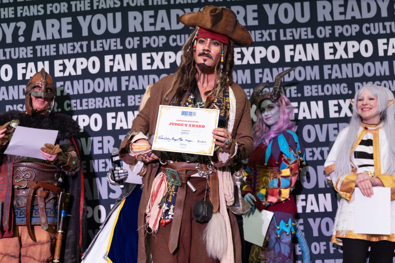 A pirate cosplayer channeling Jack Sparrow strikes a confident pose while holding a “Judge’s Award” certificate. The costume features dreadlocks, a tricorn hat, a detailed coat, and many pirate accessories. A horned fantasy character and an elf smile nearby.