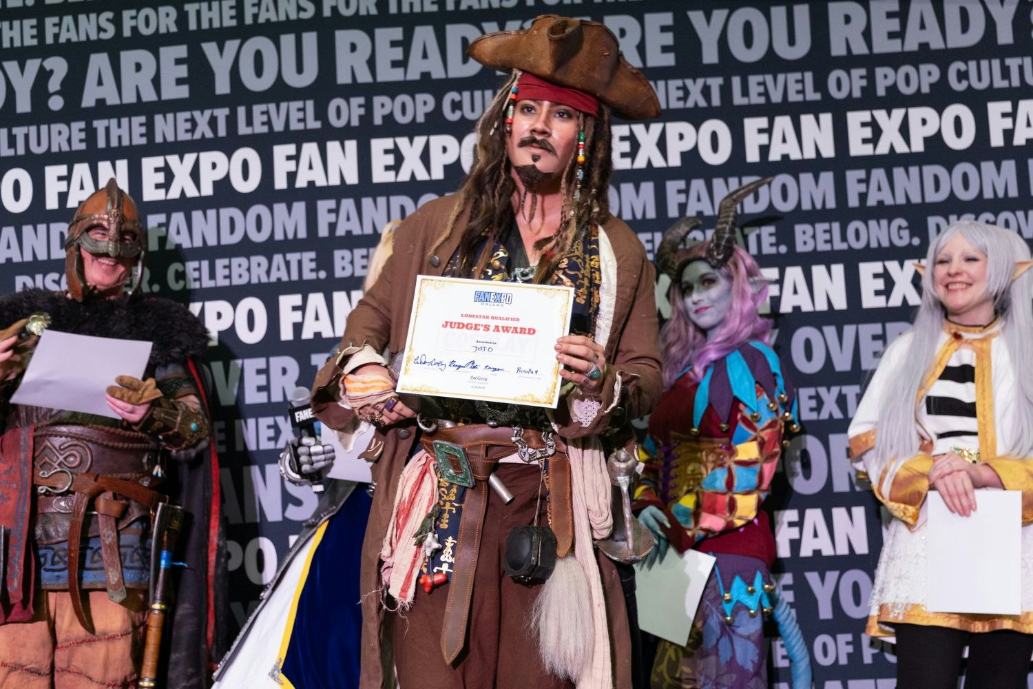 A pirate cosplayer channeling Jack Sparrow strikes a confident pose while holding a “Judge’s Award” certificate. The costume features dreadlocks, a tricorn hat, a detailed coat, and many pirate accessories. A horned fantasy character and an elf smile nearby.