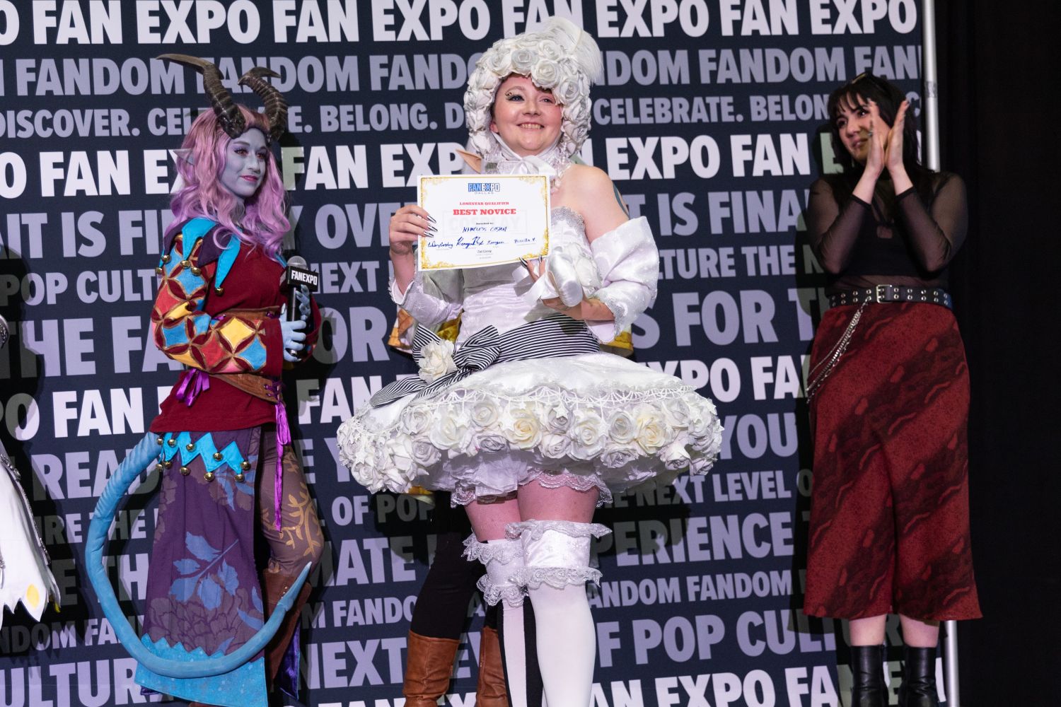 A smiling cosplayer in an all-white rococo-inspired dress embellished with roses and black stripes holds a “Best Novice” certificate. Their look includes a detailed wig with floral accents and thigh-high white boots. Fellow finalists stand to the side, applauding.