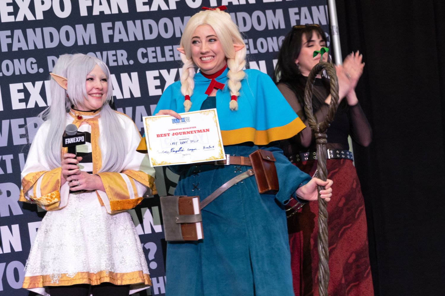 A joyful cosplayer dressed in blue and gold fantasy robes with long blonde braids and elf ears proudly holds a “Best Journeyman” certificate. A large gnarled staff leans against their shoulder. Two judges and another finalist clap enthusiastically beside them.