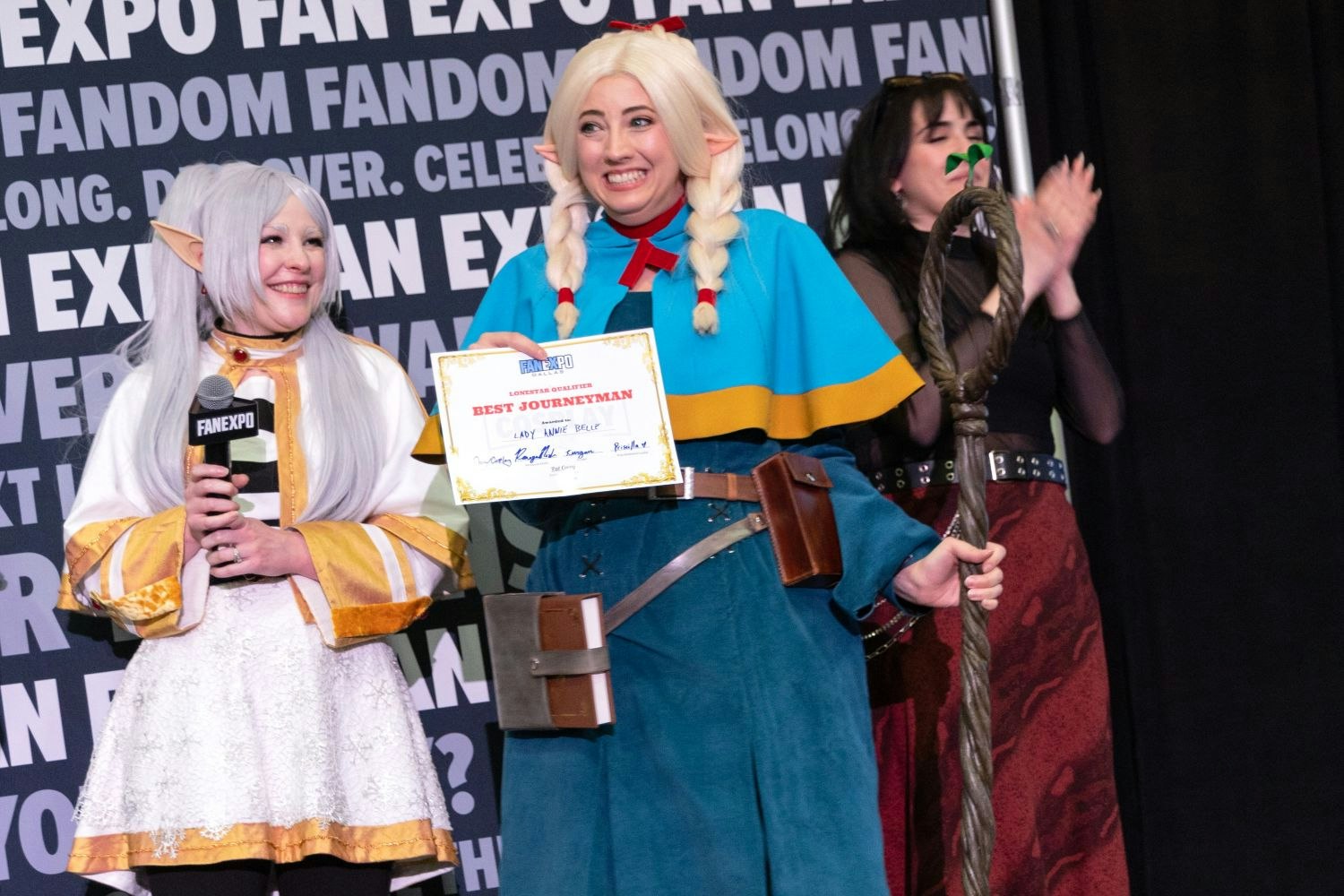 A joyful cosplayer dressed in blue and gold fantasy robes with long blonde braids and elf ears proudly holds a “Best Journeyman” certificate. A large gnarled staff leans against their shoulder. Two judges and another finalist clap enthusiastically beside them.