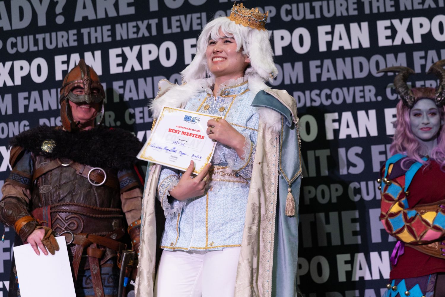 A cosplayer in an ornate royal costume smiles on stage while holding a "Best Masters" award certificate. They wear a white wig, a golden crown, and a pale blue and gold embroidered outfit with a luxurious cape. Two other detailed cosplayers flank them—one in Viking armor and another in a colorful horned look.