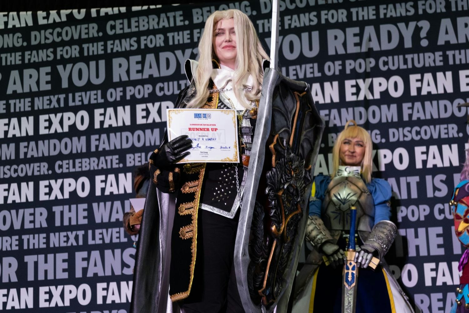 A cosplayer dressed in a long white wig and intricate black and gold vampire-style armor holds a "Runner Up" award certificate. A large sculpted pauldron shaped like a roaring beast adorns their right shoulder. Other armored and fantasy-clad contestants look on proudly in the background.