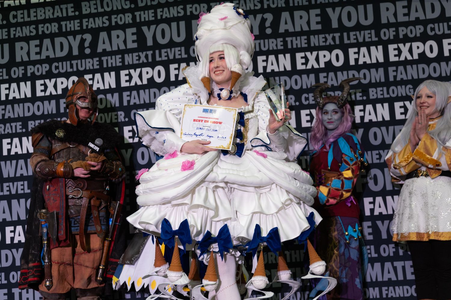 The "Best in Show" winner beams in an elaborate dessert-themed costume with giant ice cream cones and bows decorating the voluminous skirt. They hold both a trophy and certificate. Other finalists dressed in fantasy garb, including horns and elven ears, cheer from behind.