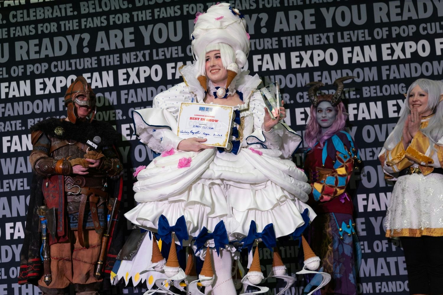 The "Best in Show" winner beams in an elaborate dessert-themed costume with giant ice cream cones and bows decorating the voluminous skirt. They hold both a trophy and certificate. Other finalists dressed in fantasy garb, including horns and elven ears, cheer from behind.