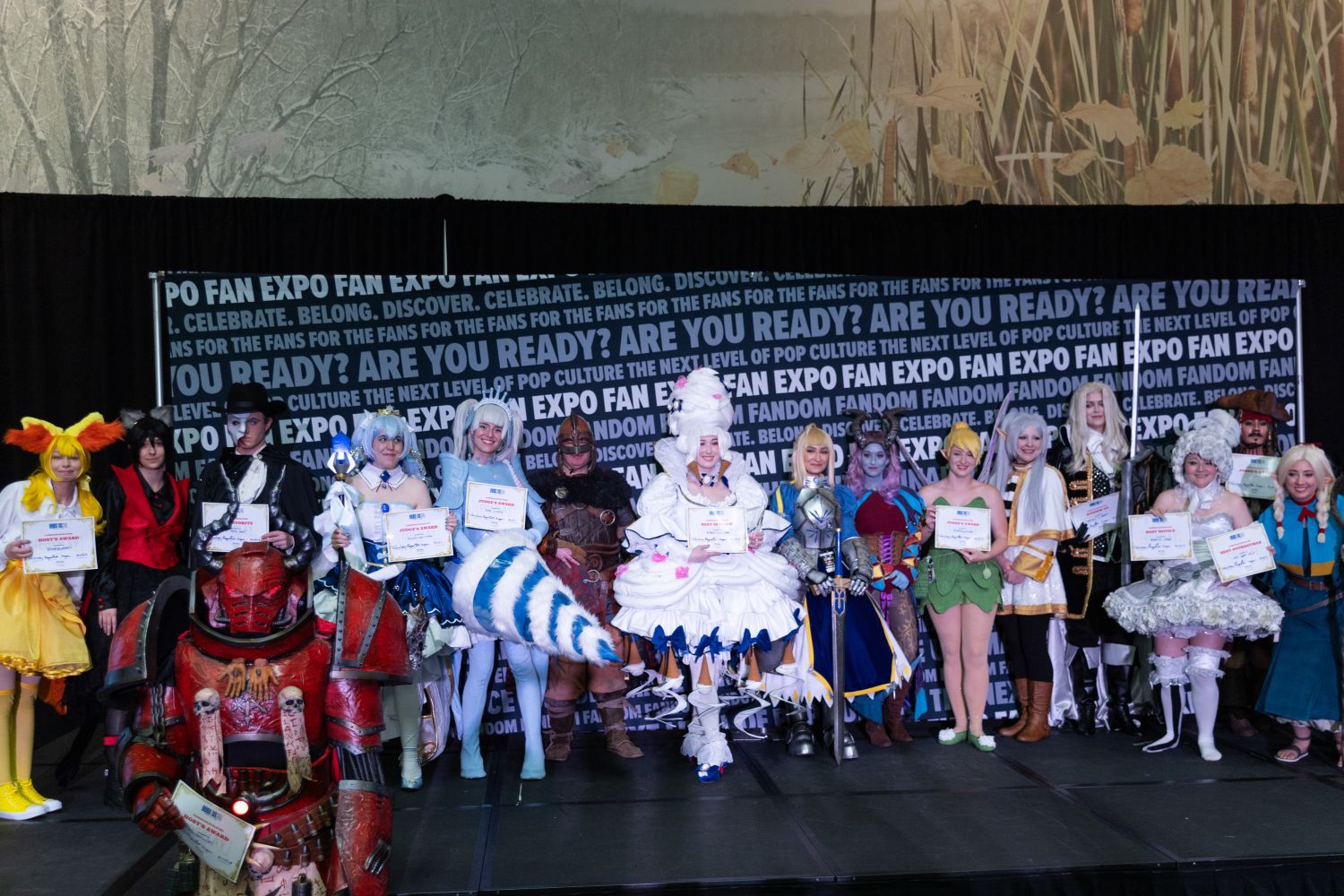 A wide group photo of all Masters of Cosplay winners and finalists posing onstage, each holding certificates. The costumes range from armored knights and fairies to towering confections and fiery mechs, showcasing a diverse range of craftsmanship and creativity.