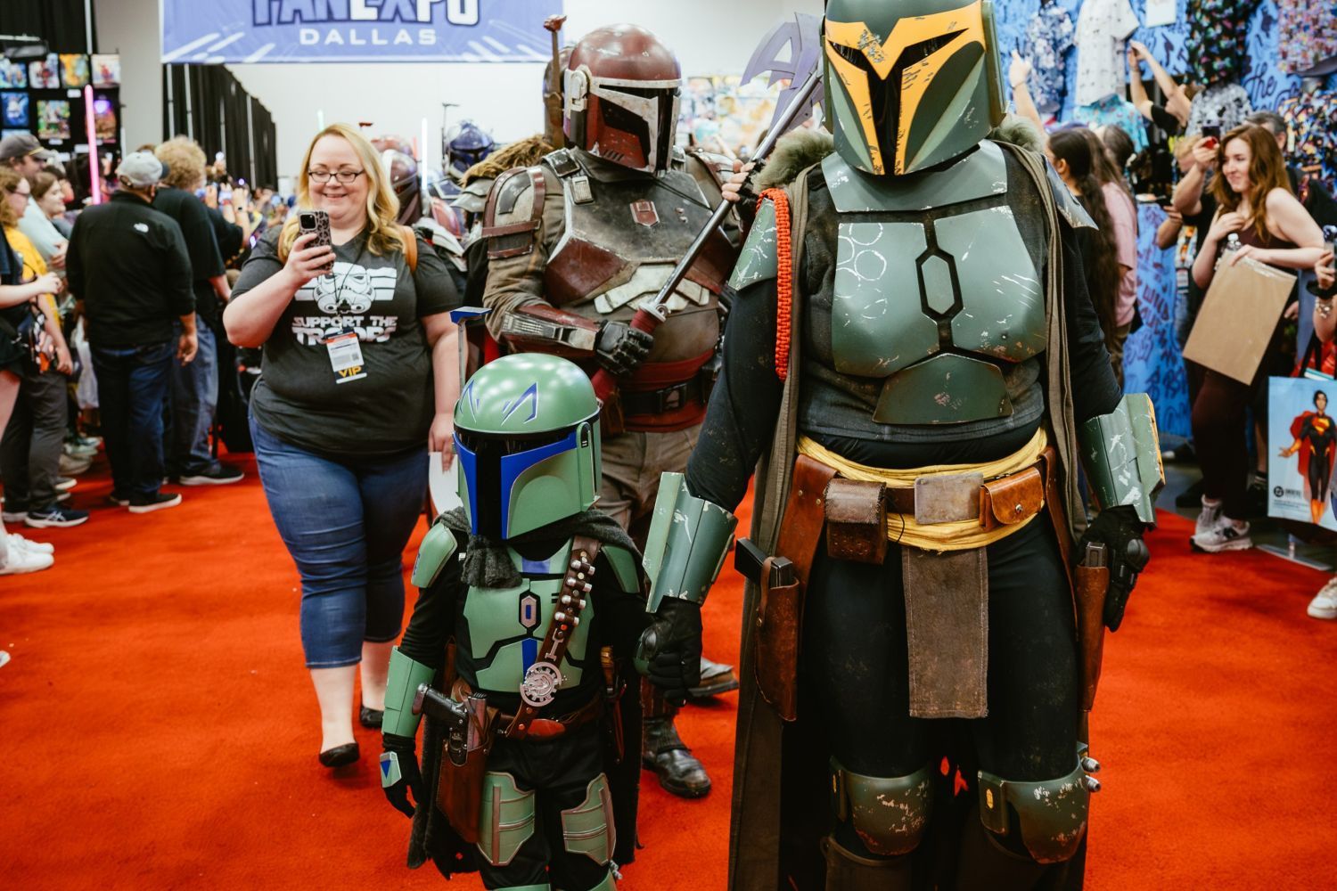 A family cosplays as Mandalorians while walking the show floor. A small child in green armor holds hands with an adult in dark green and yellow armor, with a taller Mandalorian walking behind them. A smiling woman in a "Support the Troop" shirt follows, taking a photo.