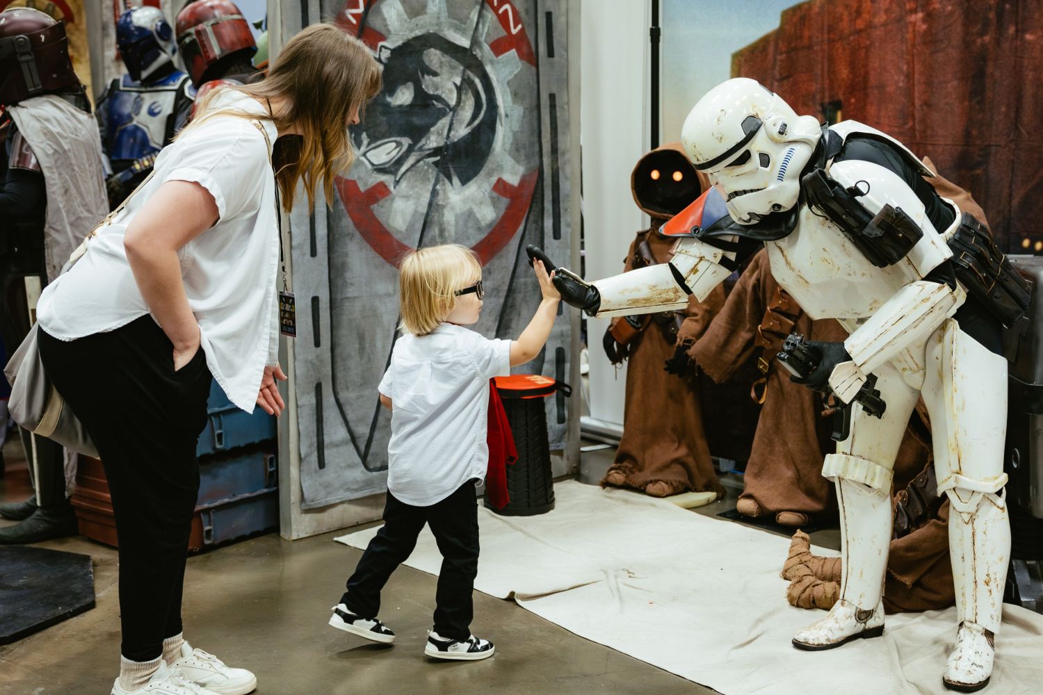A toddler in glasses high-fives a costumed Stormtrooper while their parent leans in, smiling. Jawas and Mandalorians stand in the background at a Star Wars-themed booth.