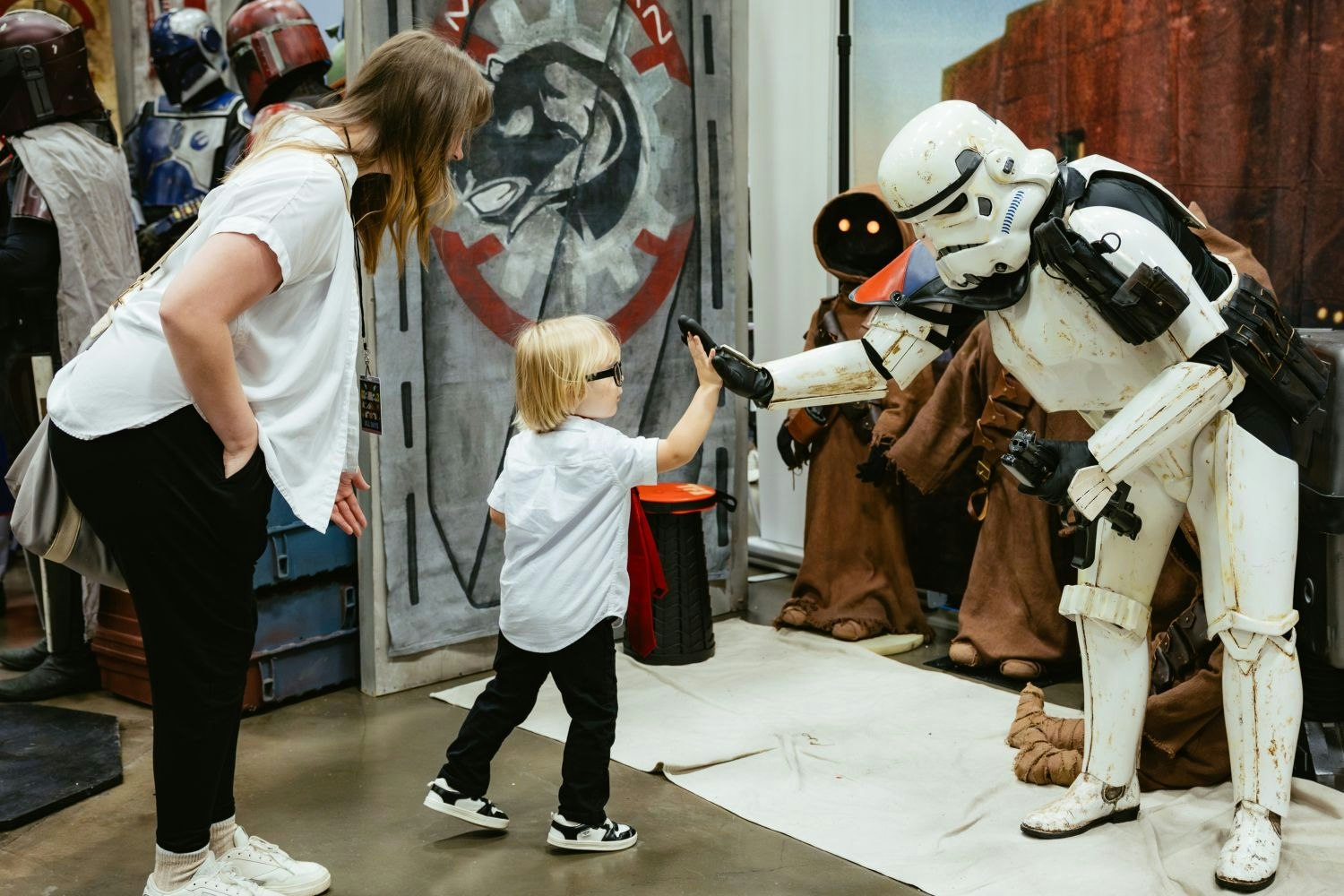 A toddler in glasses high-fives a costumed Stormtrooper while their parent leans in, smiling. Jawas and Mandalorians stand in the background at a Star Wars-themed booth.