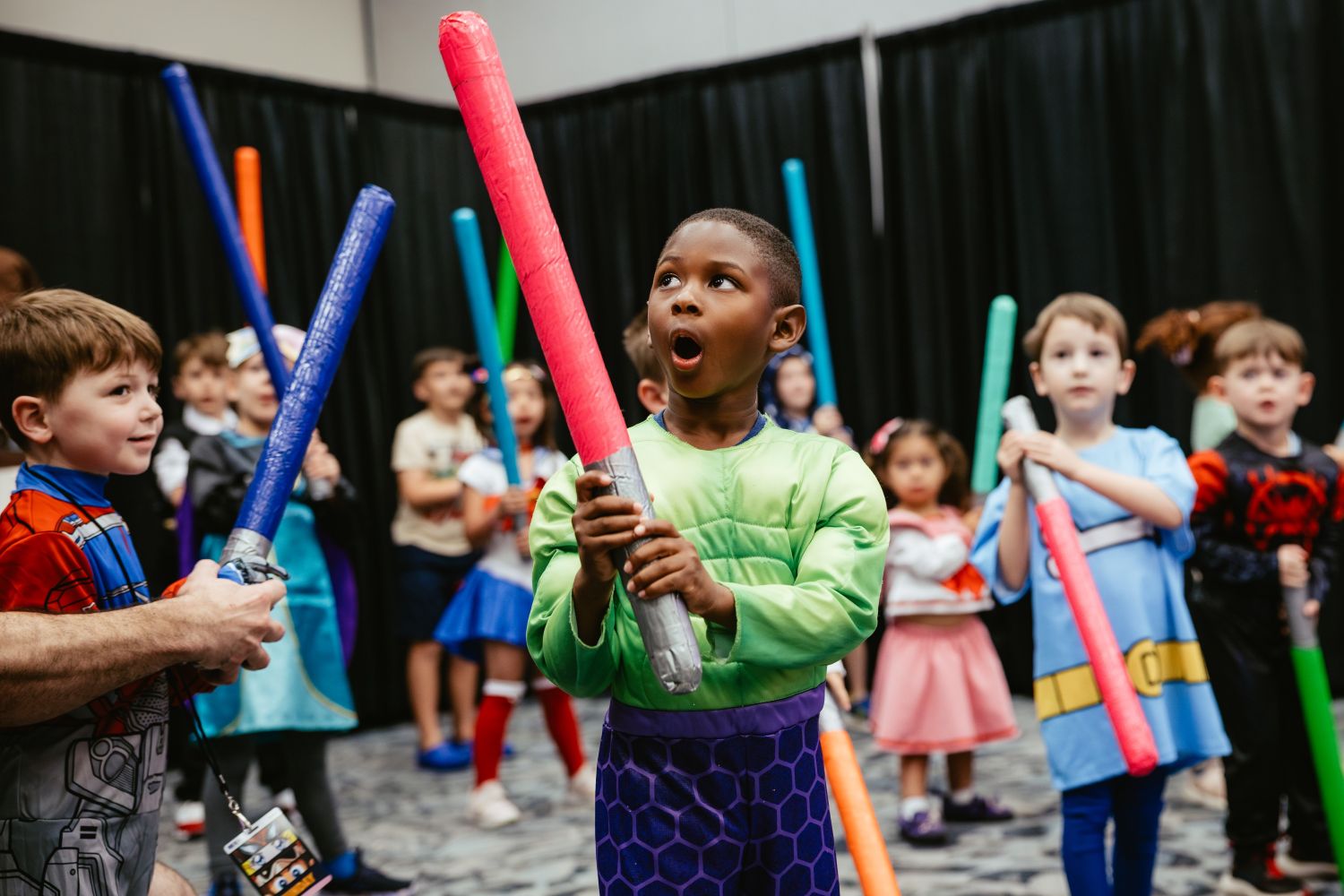 A group of young kids in costume take part in a lightsaber training activity. A child dressed as the Hulk stands front and center, jaw dropped in awe, while others around him smile and hold colorful foam sabers.