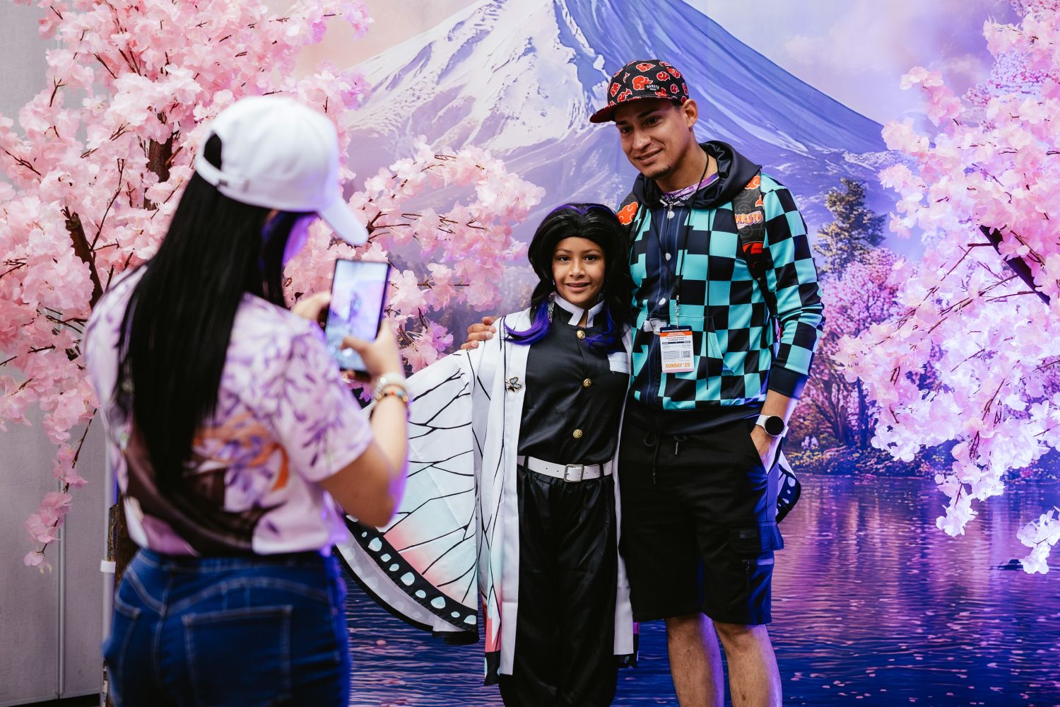 A young cosplayer dressed as Shinobu from Demon Slayer poses with a family member in front of a cherry blossom and Mt. Fuji backdrop while another adult snaps a photo on their phone.