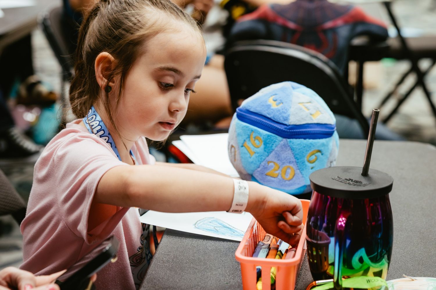 A young girl focuses intently while reaching into a bin of crayons at a craft station. A plush D20 dice sits nearby along with a colorful drink tumbler.