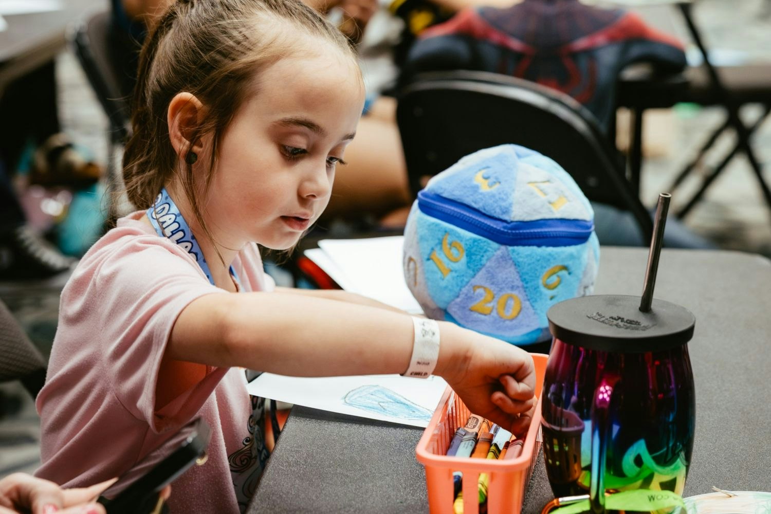 A young girl focuses intently while reaching into a bin of crayons at a craft station. A plush D20 dice sits nearby along with a colorful drink tumbler.