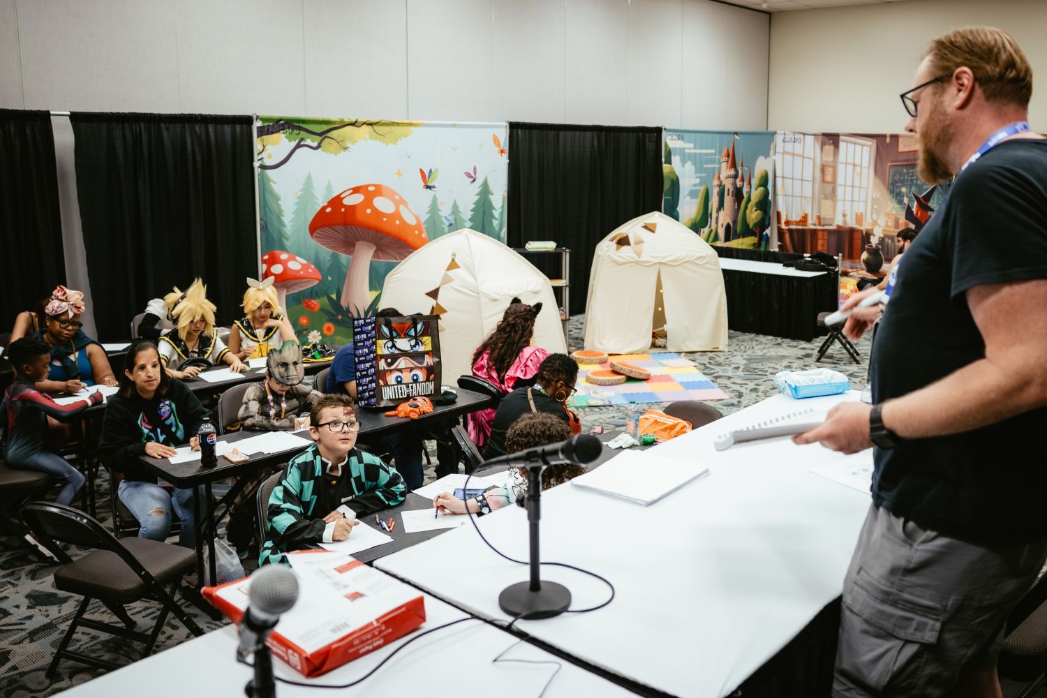 Kids and parents gather around desks in a decorated classroom setting, listening to a speaker. A fantasy forest mural and play tents decorate the back wall as children in costume work on creative projects.