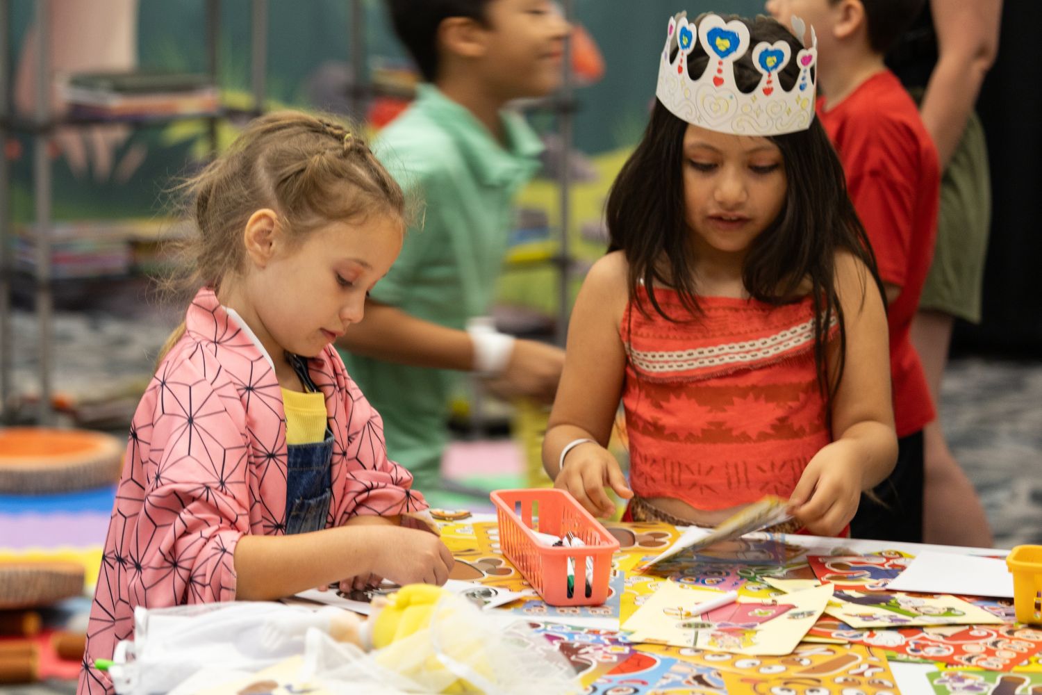 Two young girls work on a sticker activity at a colorful table. One wears a pink kimono-style jacket, and the other, dressed as Moana, wears a handmade paper crown with hearts and stars.