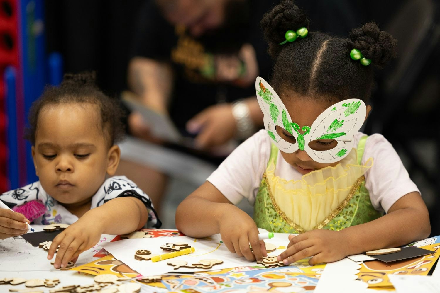 Two toddlers sit at a craft table making wooden cutout art. One wears a green fairy costume with butterfly glasses while the other concentrates on gluing pieces to her project.
