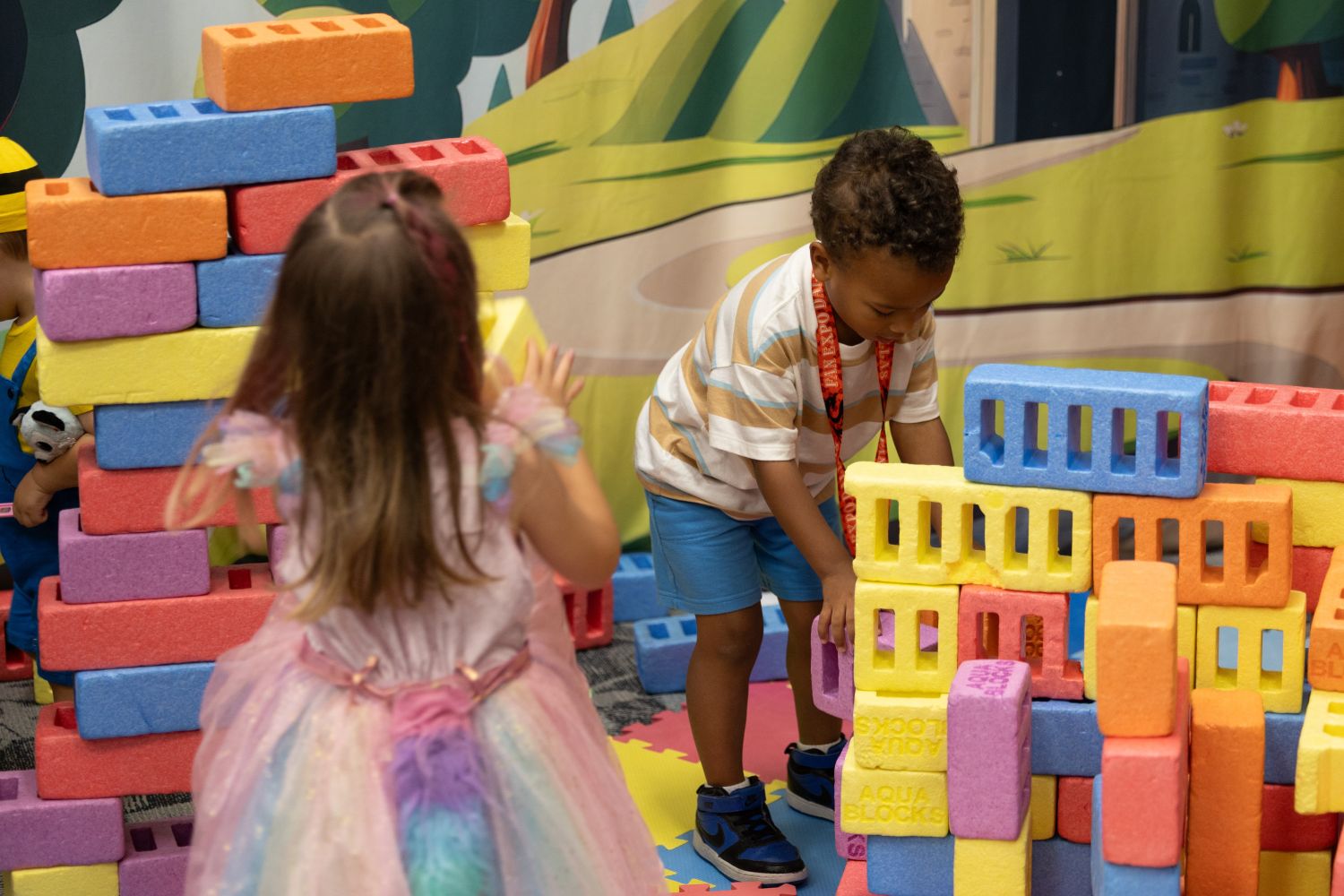 Two young kids build colorful foam block structures together in the family activity area. One wears a pastel unicorn dress, the other stacks yellow and blue blocks while wearing a striped shirt and shorts.