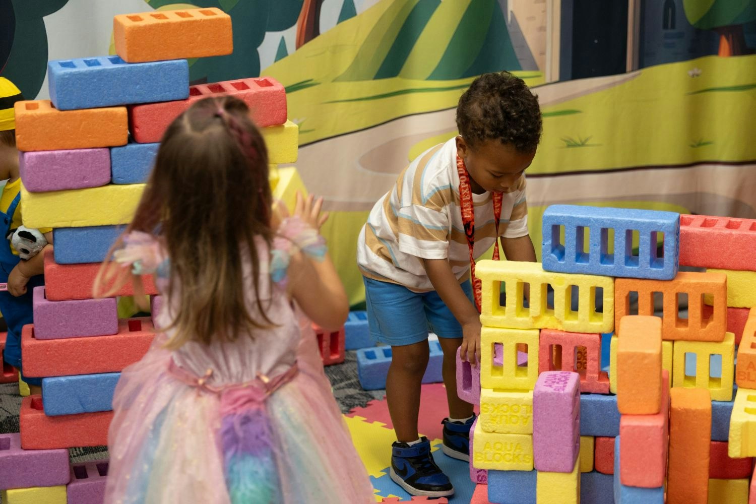 Two young kids build colorful foam block structures together in the family activity area. One wears a pastel unicorn dress, the other stacks yellow and blue blocks while wearing a striped shirt and shorts.