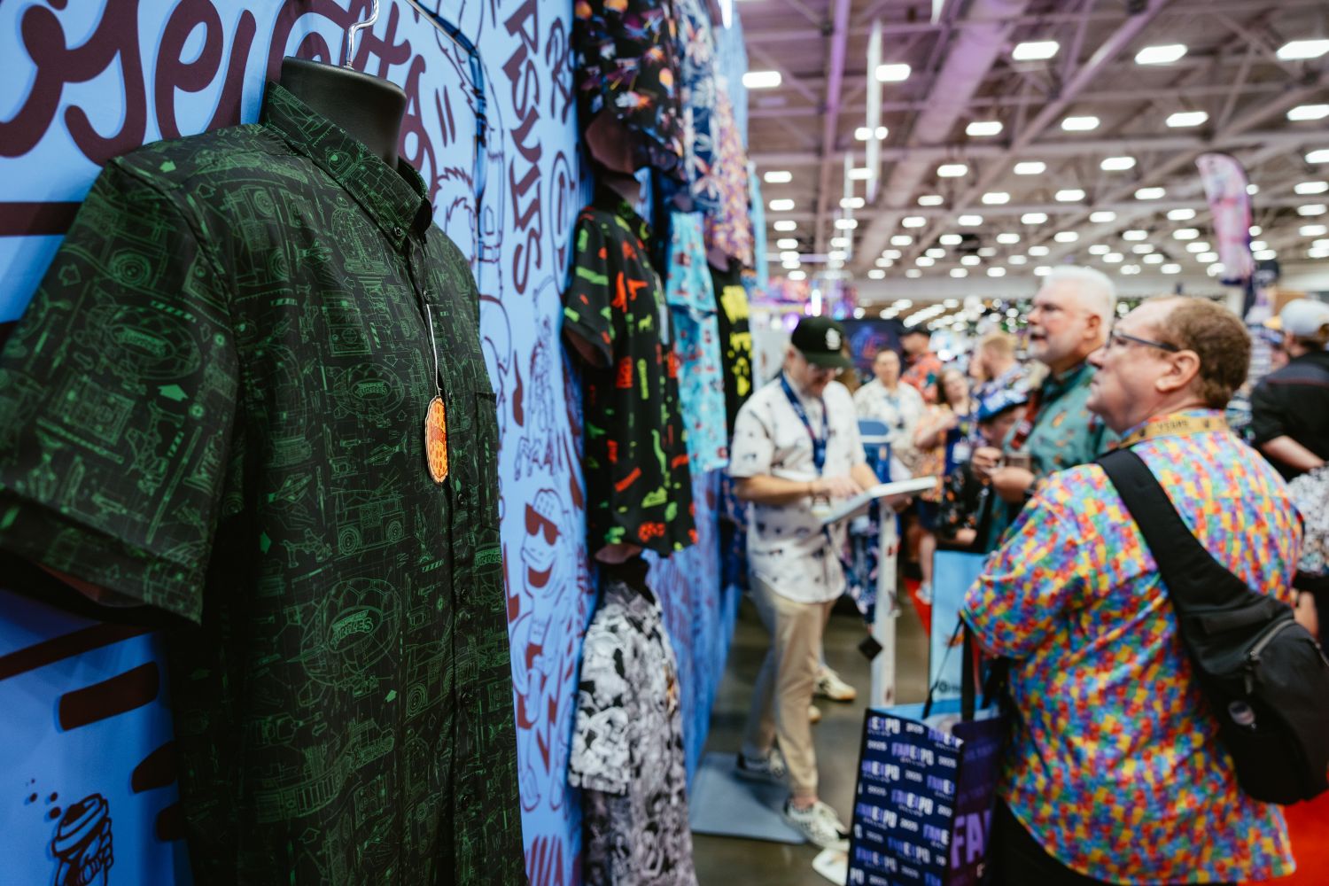 A close-up of a geeky button-up shirt covered in green schematic patterns displayed on a mannequin at a booth, with fans admiring other brightly colored shirts in the background.