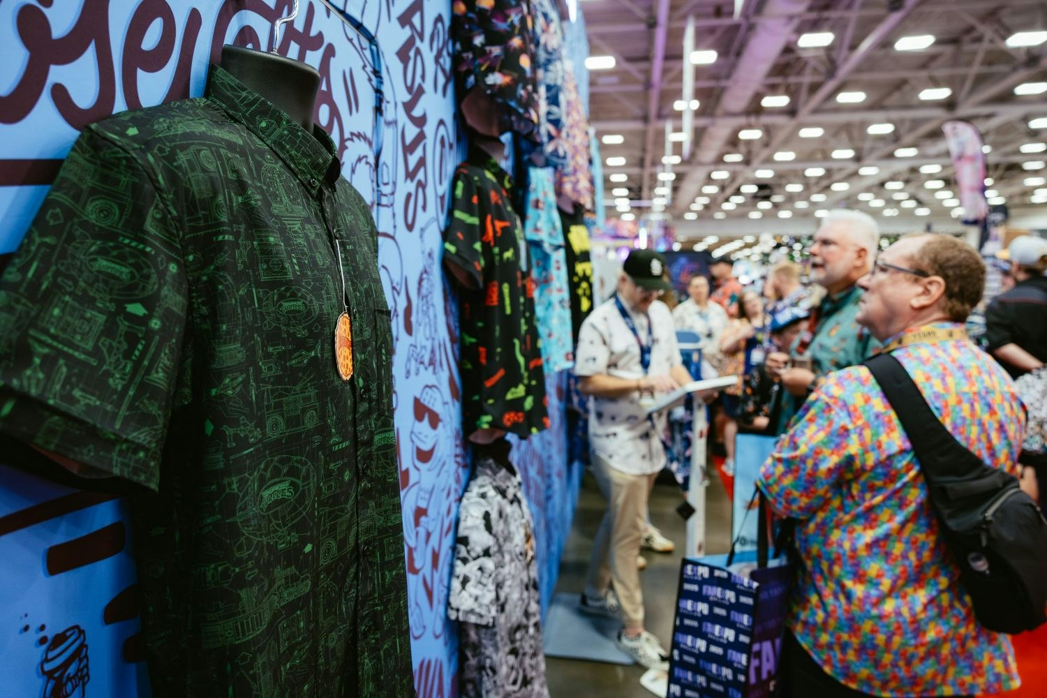 A close-up of a geeky button-up shirt covered in green schematic patterns displayed on a mannequin at a booth, with fans admiring other brightly colored shirts in the background.
