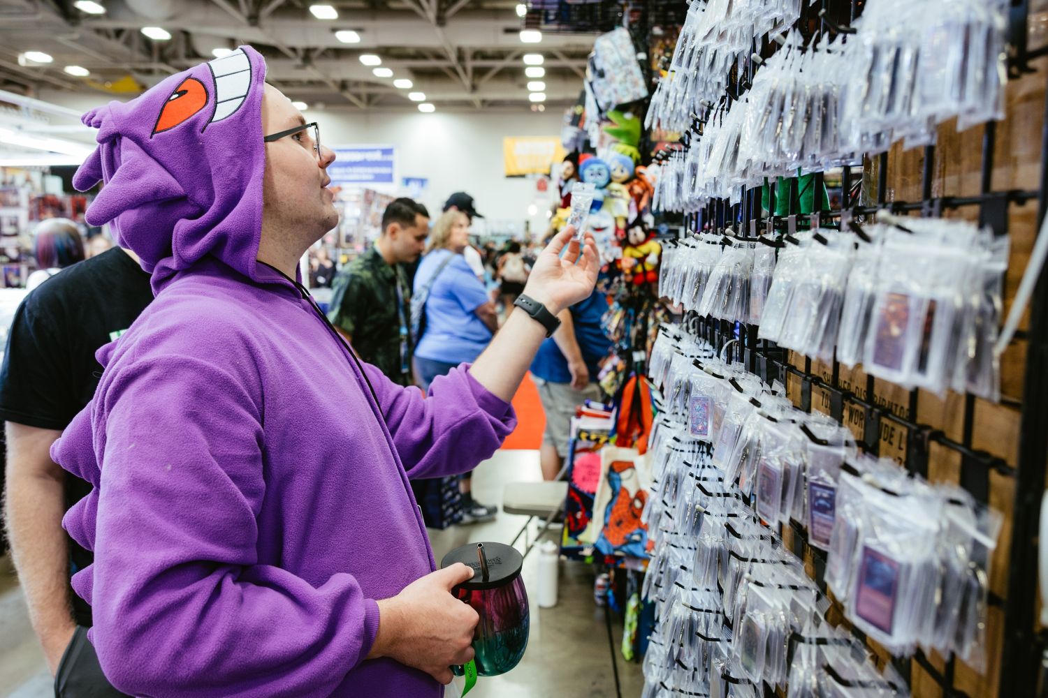 A fan wearing a purple Gengar hoodie smiles as he browses a wall of individually bagged enamel pins and keychains, holding a colorful drink tumbler in his other hand.