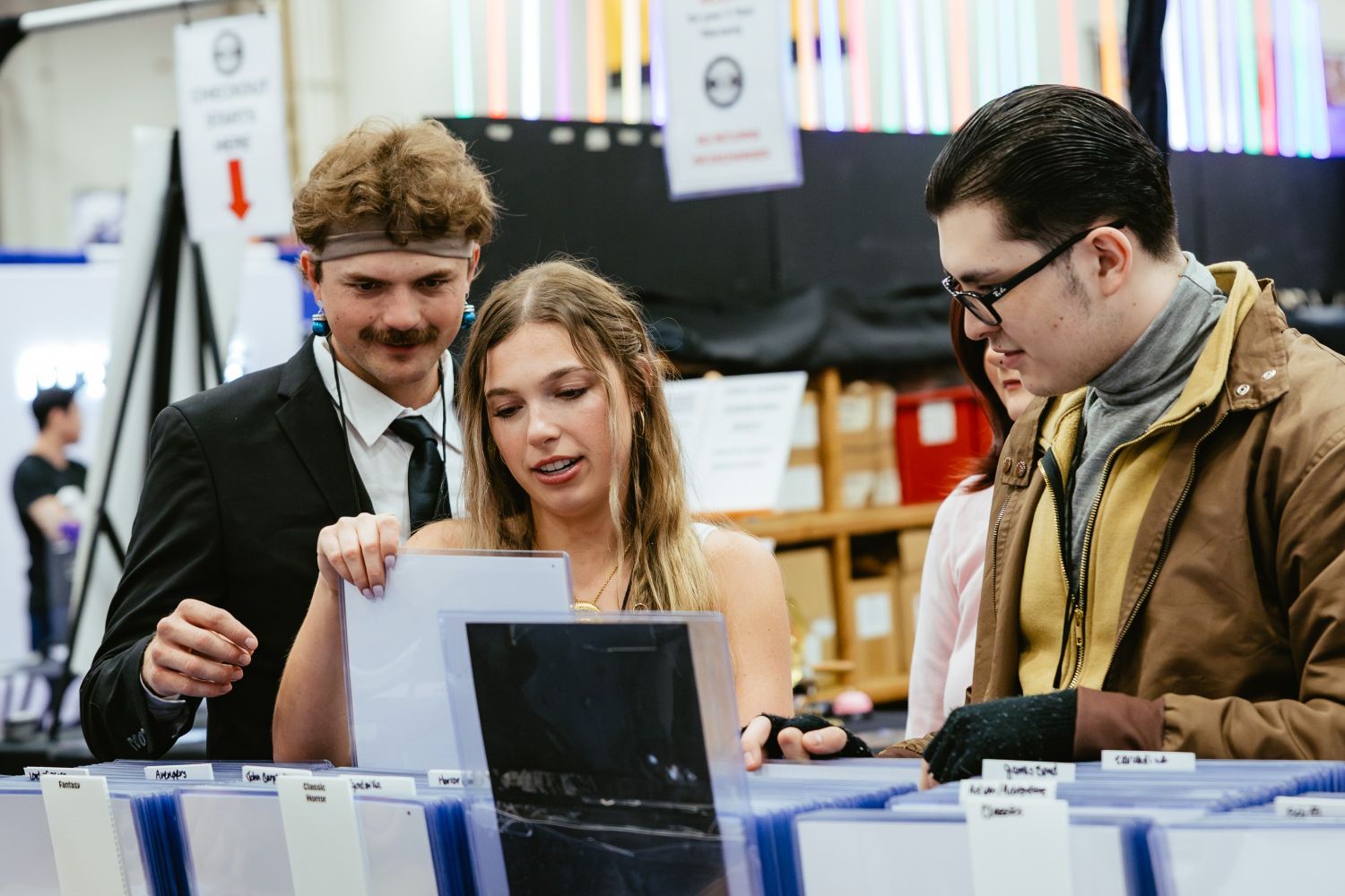Three fans browse a booth filled with protective sleeves and labeled folders for organizing comic books and artwork, discussing their finds with excitement.
