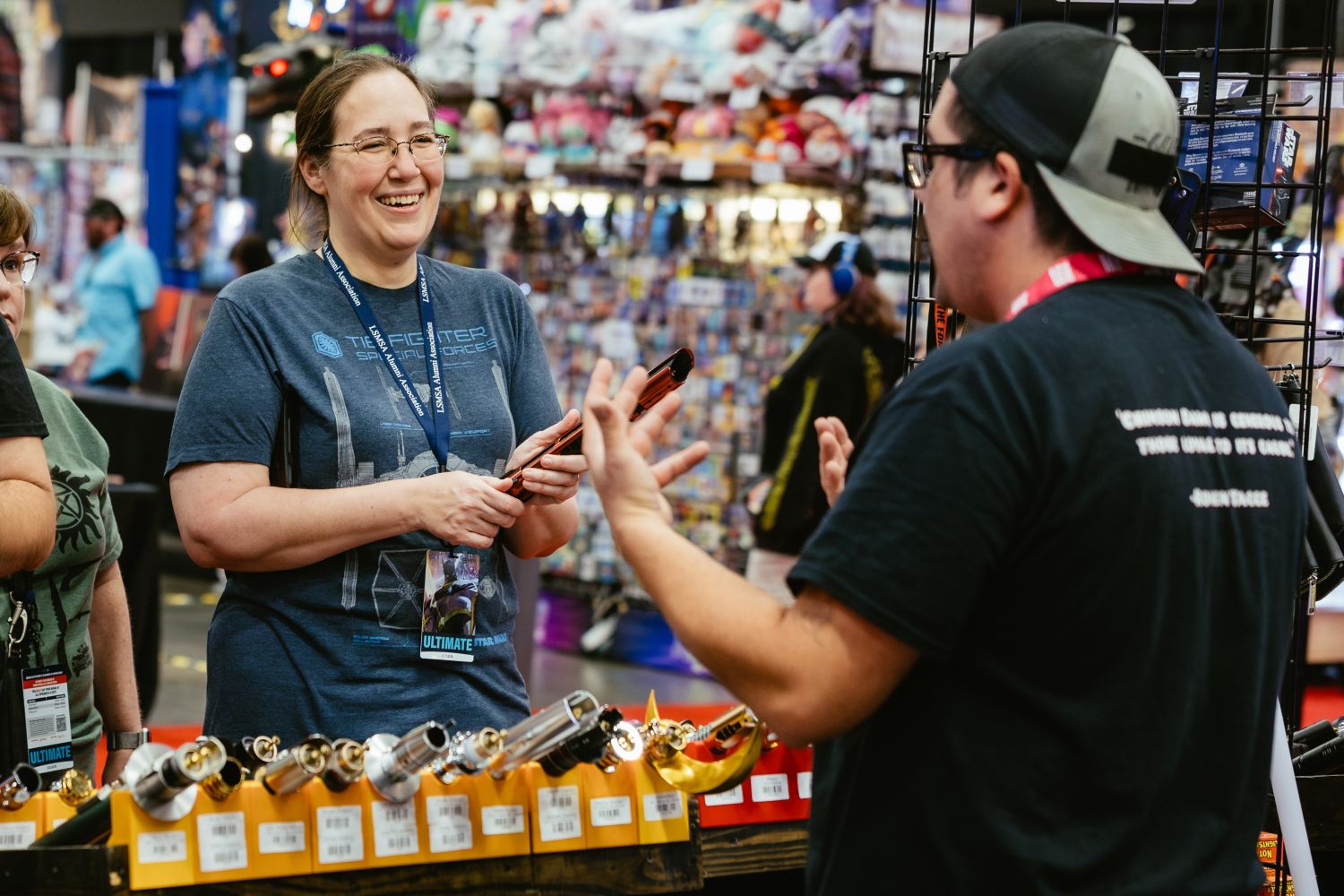 A smiling fan chats with a vendor behind a booth filled with high-end lightsaber hilts, appearing to weigh a purchase while wearing a NASA-themed t-shirt and Ultimate pass lanyard.
