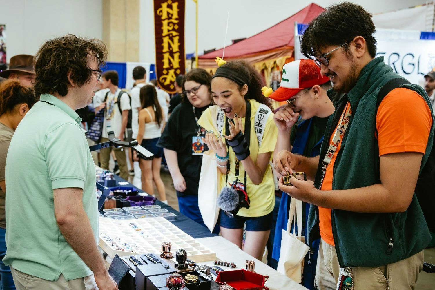 A group of friends excitedly react to custom dice and jewelry at a vendor’s table, with one attendee trying on rings and laughing while others admire geeky trinkets.