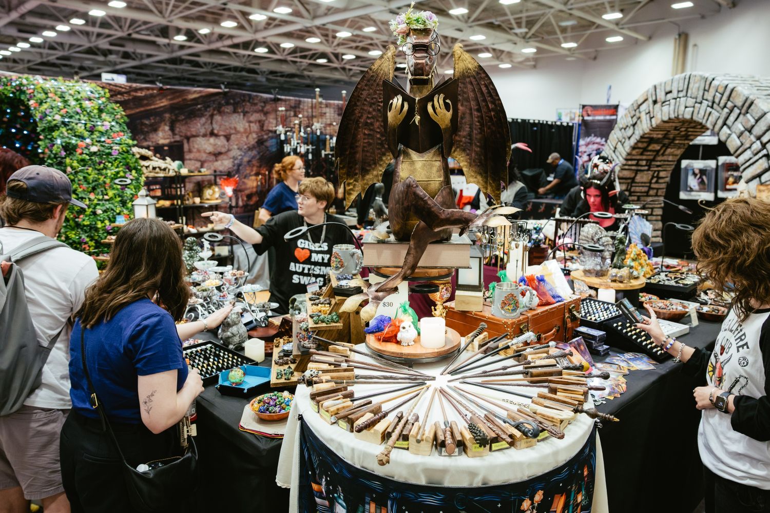 A fantasy-themed vendor booth showcases handmade wands arranged in a circular display, with shelves of crystals, jewelry, and a bronze gargoyle statue wearing glasses perched above the table.